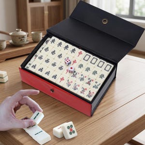 May include: A Mahjong set in a red and black case. The set includes ivory-coloured tiles with Chinese characters and symbols, dice, and a case with a snap closure. The game is displayed on a wooden table.