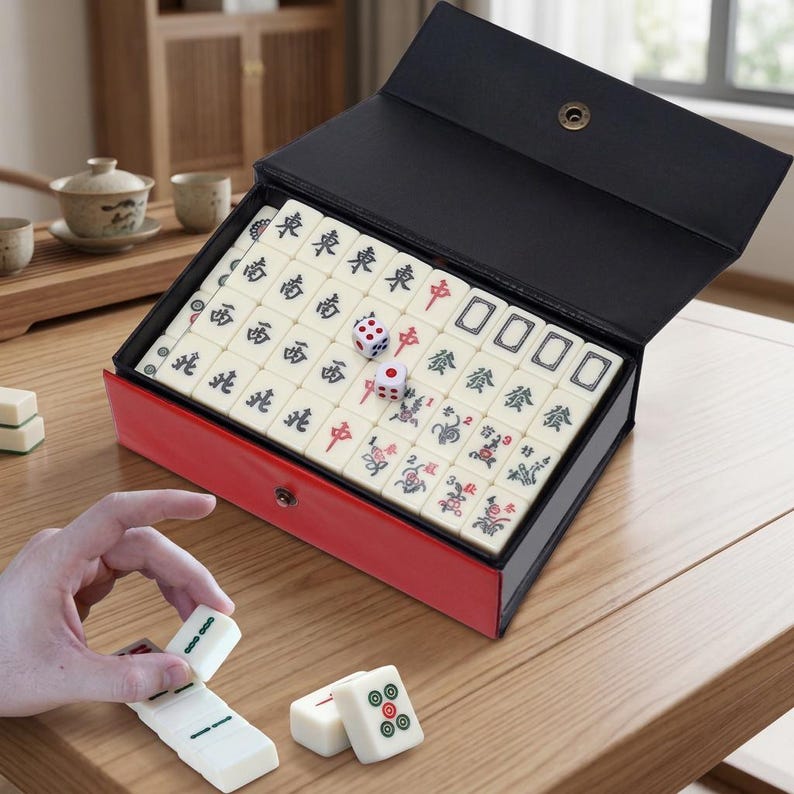 May include: A Mahjong set in a red and black case. The set includes ivory-coloured tiles with Chinese characters and symbols, dice, and a case with a snap closure. The game is displayed on a wooden table.