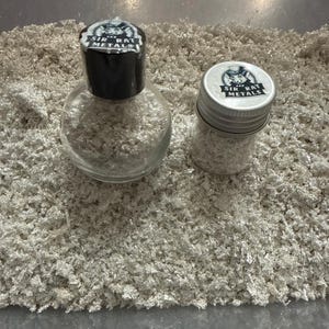 May include: Two glass jars filled with silver flakes, one with a black cap and the other with a silver lid. Both jars have a label that reads "SIR RAT METALS" with a cartoon rat. The jars sit on a bed of silver flakes.