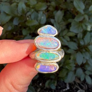 May include: A silver ring with five stacked, oval opals. The opals display blue, orange, green, and purple hues. The ring is held up against a backdrop of green foliage.