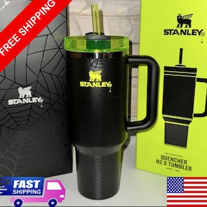 May include: Black Stanley Quencher H2.0 Tumbler with a green lid and straw. The tumbler has a handle and the Stanley logo in yellow. The packaging is visible in the background.