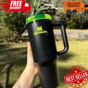 May include: Black Stanley tumbler with a green lid and handle. The Stanley logo is in yellow. The tumbler is held in front of a blurred green background. The words "Free Delivery" and "Delivery Service" are visible.