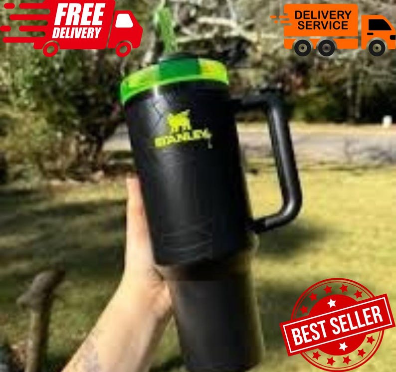 May include: Black Stanley tumbler with a green lid and handle. The Stanley logo is in yellow. The tumbler is held in front of a blurred green background. The words "Free Delivery" and "Delivery Service" are visible.