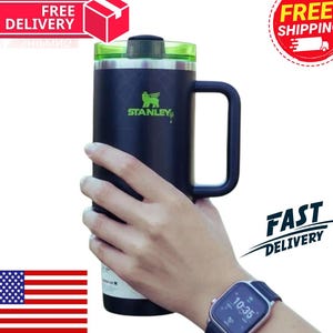 May include: A black Stanley travel mug with a green lid and handle. The mug features the Stanley logo in green and the text "STANLEY". The mug is held by a person.