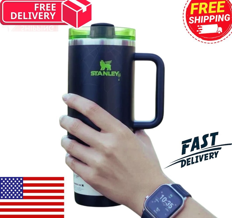 May include: A black Stanley travel mug with a green lid and handle. The mug features the Stanley logo in green and the text "STANLEY". The mug is held by a person.