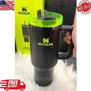 May include: Black Stanley tumbler with a green lid and handle. The tumbler has the Stanley logo in yellow. A yellow box with the Stanley logo is in the background. The tumbler is held by a person.