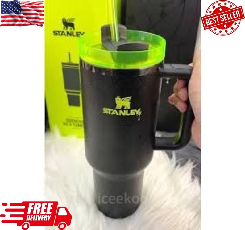 May include: Black Stanley tumbler with a green lid and handle. The tumbler has the Stanley logo in yellow. A yellow box with the Stanley logo is in the background. The tumbler is held by a person.