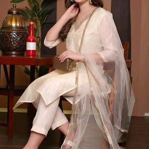 May include: An off-white salwar kameez outfit with embroidery and a matching dupatta. The ensemble includes a V-neck top and white sandals. A small, brown clutch is held. The background features a table, a decorative lamp, and a plant.