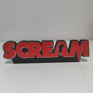 Op de afbeelding: Een driedimensionaal bord met het woord SCREAM in dikke rode hoofdletters met een zwarte omlijning. De letter A bevat een zwart silhouet van een rennend figuur. Het bord heeft een stevige zwarte basis en staat rechtop.