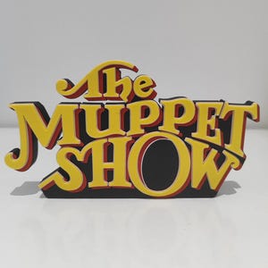 Puede incluir: Un letrero tridimensional que presenta el texto The Muppet Show en letras amarillas con acentos en capas de color rojo y negro. Las letras están estilizadas con una fuente curva y negrita, creando un efecto de capas sobre el fondo negro.