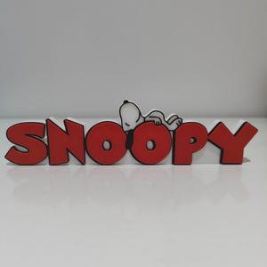 Può includere: Un'insegna decorativa con la parola SNOOPY in grassetto rosso a lettere maiuscole con un bordo bianco. Il personaggio Snoopy è raffigurato in bianco e nero, sdraiato e appoggiato sulle due lettere O al centro dell'insegna.