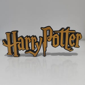 Puede incluir: Un letrero tridimensional que presenta el texto Harry Potter en una fuente estilizada. Las letras son de color amarillo mostaza con un borde negro grueso, creando un efecto de capas que permite que el letrero se mantenga en pie.