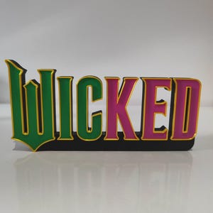 Può includere: Un'insegna tridimensionale che mostra la parola WICKED in un carattere stilizzato. Le lettere W, I, C e K sono verdi con bordi dorati, mentre le lettere E e D sono rosa con bordi dorati. Il tutto è montato su una base nera solida.