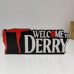 Könnte beinhalten: Ein schwarzes rechteckiges Schild mit dem roten Text IT und dem weißen Text WELCOME TO DERRY. Der Buchstabe O in WELCOME ist durch eine rote Ballonform ersetzt. Das Schild hat eine matte Oberfläche und steht aufrecht auf einer flachen Fläche.