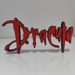 Könnte beinhalten: Ein freistehendes Dekoschild mit dem Wort Dracula in einer stilisierten, tropfenden Schriftart. Die Buchstaben sind leuchtend rot mit einer dicken schwarzen Umrandung, was einen geschichteten, dreidimensionalen Effekt erzeugt, der aufrecht steht.