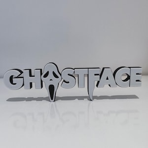 May include: A three-dimensional sign featuring the word GHOSTFACE in bold, white, block lettering with black outlines. The letter O is replaced by a white mask with black eye and mouth cutouts, and the letter T features a downward-pointing extension.