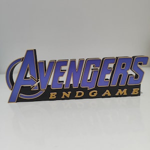 Può includere: Un'insegna tridimensionale che presenta il logo Avengers in viola con bordi dorati, posizionata sopra la parola ENDGAME in lettere dorate su una base nera. Il design mostra l'iconico titolo del film con un aspetto stratificato e in rilievo.