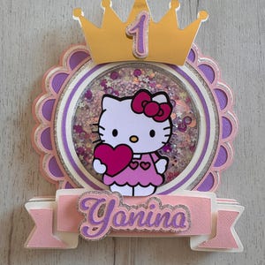 Puede incluir: Adorno para tarta en capas con Hello Kitty sosteniendo un corazón, dentro de un círculo transparente lleno de confeti. El diseño incluye una corona dorada con el número 1 y una pancarta que dice "Yanina" en letras moradas brillantes. La combinación de colores es principalmente rosa y morado.