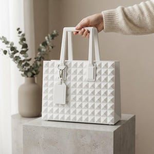 Puede incluir: Un bolso de mano rectangular de color blanco s&oacute;lido con una superficie exterior texturizada de pir&aacute;mides geom&eacute;tricas. El bolso incluye dos asas r&iacute;gidas verticales y una etiqueta rectangular a juego sujeta a una anilla de metal plateado.