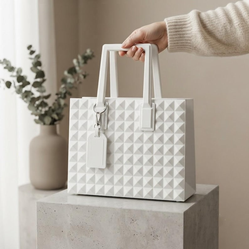Puede incluir: Un bolso de mano rectangular de color blanco s&oacute;lido con una superficie exterior texturizada de pir&aacute;mides geom&eacute;tricas. El bolso incluye dos asas r&iacute;gidas verticales y una etiqueta rectangular a juego sujeta a una anilla de metal plateado.