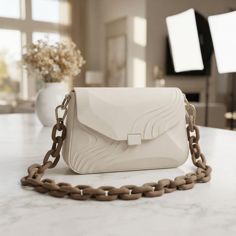 Puede incluir: Un bolso de hombro color crema con una solapa estructurada que presenta l&iacute;neas onduladas en relieve y un cierre rectangular central. El bolso incluye una correa de cadena gruesa de color marr&oacute;n con eslabones entrelazados y herrajes dorados.