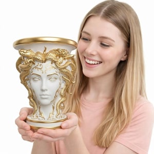 Puede incluir: Un jarrón blanco y dorado con forma de cabeza de Medusa. El jarrón presenta una representación detallada del rostro de Medusa con serpientes y detalles dorados. El jarrón tiene una parte superior ancha con borde dorado y una base decorativa.