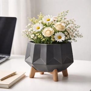 Op de afbeelding: Een donkergrijze keramische plantenbak met een geometrisch gefacetteerd oppervlak, ondersteund door vier lichtgekleurde houten poten. De plantenbak is gevuld met een boeket witte margrieten, crèmekleurige rozen en kleine gele bloemen.