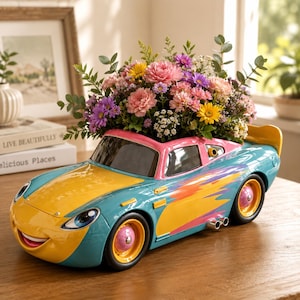 May include: A decorative planter shaped like a cartoon race car with a teal body, yellow hood, and pink wheels. The car features a smiling face on the front bumper and a vibrant orange flame graphic on the side. It is filled with a colorful bouquet.