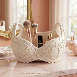 Może przedstawiać: Kremowy organizer do makijażu w kształcie biustonosza z uchwytami i misternymi koronkowymi detalami. Organizer zawiera pędzle do makijażu, szminkę i butelki perfum. Tekst "LACE & PEARLS MAKEUP ORGANIZER" znajduje się na dole.