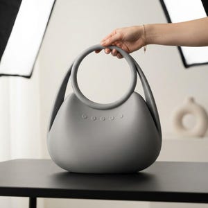 Puede incluir: Un bolso de mano gris mate y liso con un asa superior circular y un cuerpo curvo en forma de semic&iacute;rculo. La parte frontal del bolso presenta una fila horizontal de cinco peque&ntilde;os detalles circulares en relieve bajo el asa.