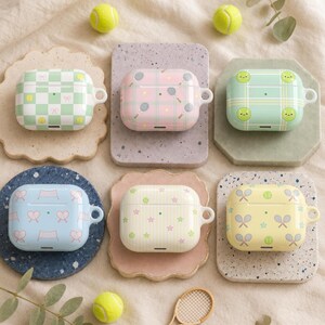 May include: Six rectangular protective cases for wireless earbuds, each featuring distinct tennis-themed patterns including checkered, plaid, and striped designs with tennis rackets, balls, and stars in pastel shades of green, pink, blue, and yellow.