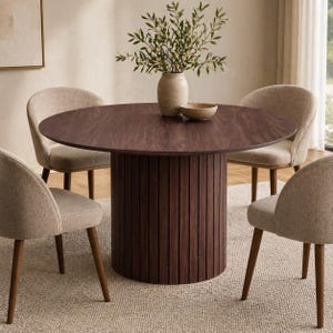 Può includere: Un tavolo da pranzo rotondo con finitura in legno scuro e base cilindrica con scanalature verticali. Quattro sedie beige imbottite con gambe in legno circondano il tavolo. Un vaso con verde e una piccola ciotola sono sul tavolo.