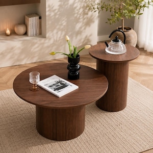 May include: Two dark brown wooden coffee tables, one round and large, the other smaller. A glass teapot sits on the smaller table. A vase of flowers and a magazine are on the larger table. The tables are on a beige rug.