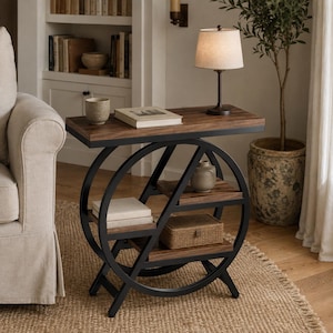 Puede incluir: Una mesa auxiliar con una superficie rectangular de madera y dos estantes inferiores de madera, sostenidos por un marco circular de metal negro. La mesa sostiene una lámpara pequeña, un libro, un cuenco de cerámica, un jarrón y una caja tejida.