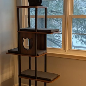 May include: A multi-level cat tree featuring dark wood platforms and black support poles. The structure includes a central enclosed box with a cat-shaped cutout, multiple rectangular gray cushions on each level, and a black cat sitting on top.