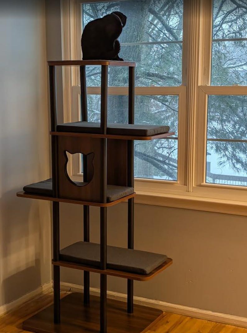 May include: A multi-level cat tree featuring dark wood platforms and black support poles. The structure includes a central enclosed box with a cat-shaped cutout, multiple rectangular gray cushions on each level, and a black cat sitting on top.