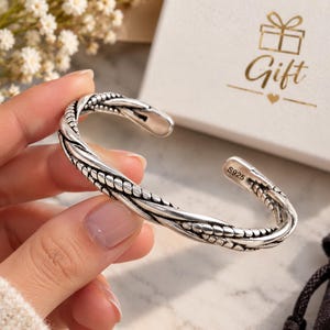 Puede incluir: Una pulsera abierta de plata esterlina con un diseño detallado de cuerda trenzada y un acabado oxidado que resalta la textura. La banda interior está claramente marcada con el sello S925, lo que indica su composición de plata.