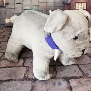 Op de afbeelding: Een zachtgrijze bulldog-knuffel met een blauwe halsband en een witte kegel. Het speelgoed staat op een bakstenen oppervlak, met een achtergrond van een stadsgezicht onder een sterrenhemel.