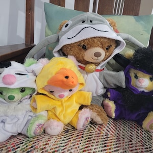 May include: A group of teddy bear plush toys, each wearing a different animal costume. The costumes include a frog, duck, cow, and a horned creature, in colours such as white, yellow, green, and purple. The toys are arranged on a patterned surface.