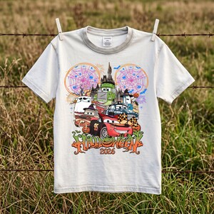 May include: A light gray short-sleeve t-shirt featuring a colorful graphic of animated cars in front of a haunted castle. The design includes spider webs, pumpkins, and the text "HALLOWEEN 2026" in a stylized orange font at the bottom.