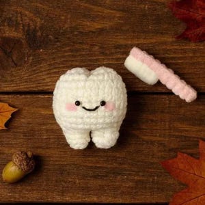 May include: A white crochet tooth with a smiling face and pink cheeks, alongside a pink and white crochet toothbrush. The tooth sits on a wooden surface with autumn leaves and an acorn.