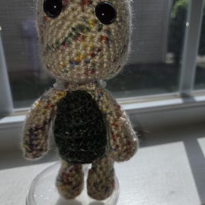 May include: A handmade crochet doll with a speckled beige head and limbs, large black button eyes, and a dark green torso. The doll is standing on a clear plastic base, showcasing its unique, handcrafted design.