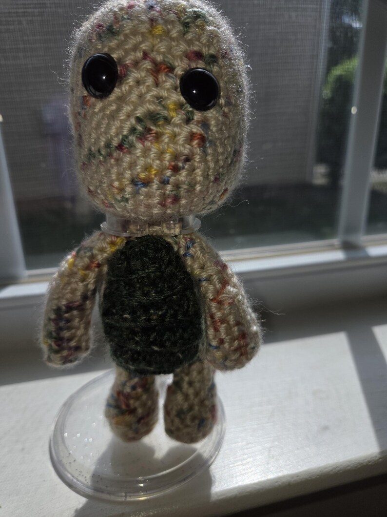 May include: A handmade crochet doll with a speckled beige head and limbs, large black button eyes, and a dark green torso. The doll is standing on a clear plastic base, showcasing its unique, handcrafted design.