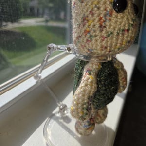 May include: A handmade crochet doll with a speckled beige head and large black eyes. The doll has a green and beige body and is mounted on a clear plastic stand. The doll is positioned in front of a window.