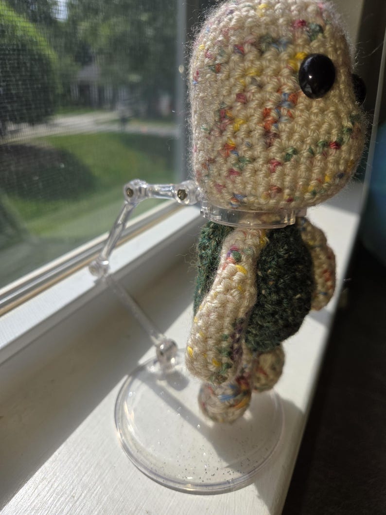 May include: A handmade crochet doll with a speckled beige head and large black eyes. The doll has a green and beige body and is mounted on a clear plastic stand. The doll is positioned in front of a window.