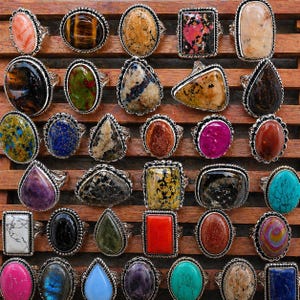 Op de afbeelding: Een collectie van dertig ringen met diverse gepolijste edelstenen in verschillende vormen, waaronder ovaal, rechthoekig, peervormig en vierkant. Elke steen is gezet in een zilverkleurige metalen zetting met decoratieve kralenranden.