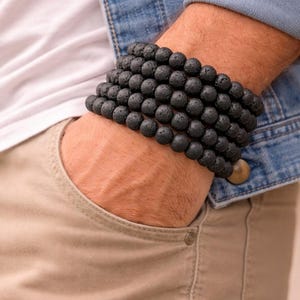 Può includere: Bracciale con perline in pietra lavica nera. Il bracciale è composto da più fili di perline nere rotonde e porose. Il bracciale è indossato al polso. La persona indossa una maglietta bianca, una giacca di jeans e pantaloni color cachi.