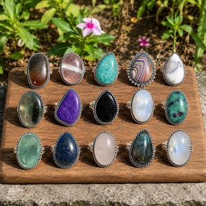 Puede incluir: Una colección de anillos de piedras preciosas expuestos sobre una superficie de madera. Los anillos presentan varias piedras de forma ovalada y de lágrima en colores como verde, morado, negro y azul, engastadas en metal plateado. El fondo incluye vegetación y algunas flores rosas.