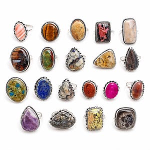 May include: A collection of twenty gemstone rings with silver-colored bands. The rings feature various shapes and colors of stones, including pink, brown, blue, and green. The stones are set in ornate, detailed settings.