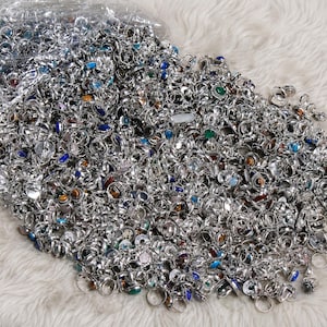 May include: A large collection of silver-toned metal rings featuring various inlaid gemstones in colors including blue, green, brown, and white. The rings are displayed in a pile, with some still contained within a clear plastic bag.